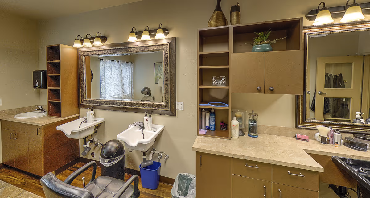 Interior view of a salon or grooming area with two white washbasins, a large mirror above them, and a salon chair in front. The room has wooden cabinets, shelves with various hair and beauty products, and two wall-mounted light fixtures above the mirrors. There is a window with curtains reflected in the mirror.