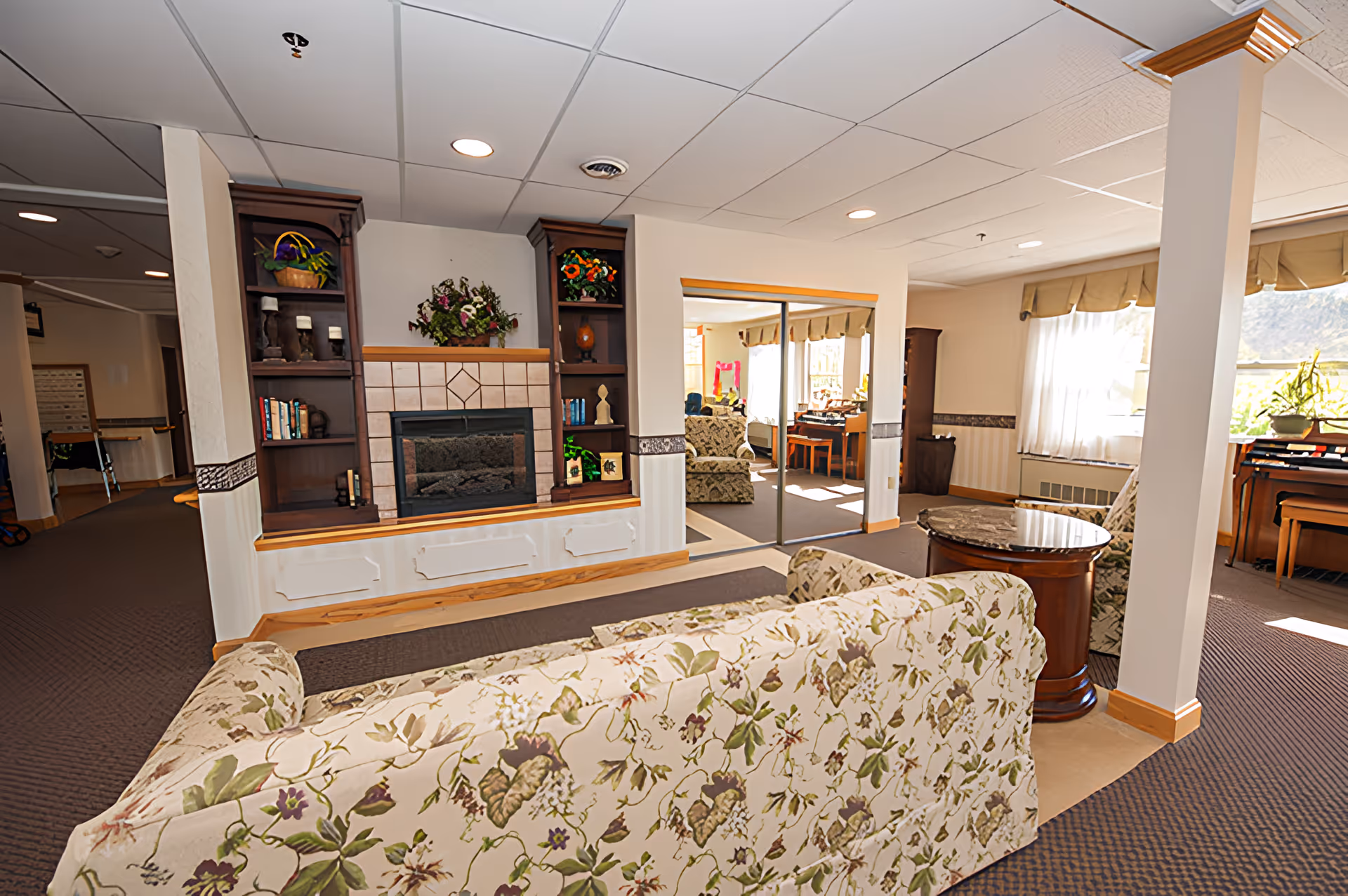 Sunlit communal living room with floral sofas, a fireplace flanked by built-in shelves, and seating near windows.