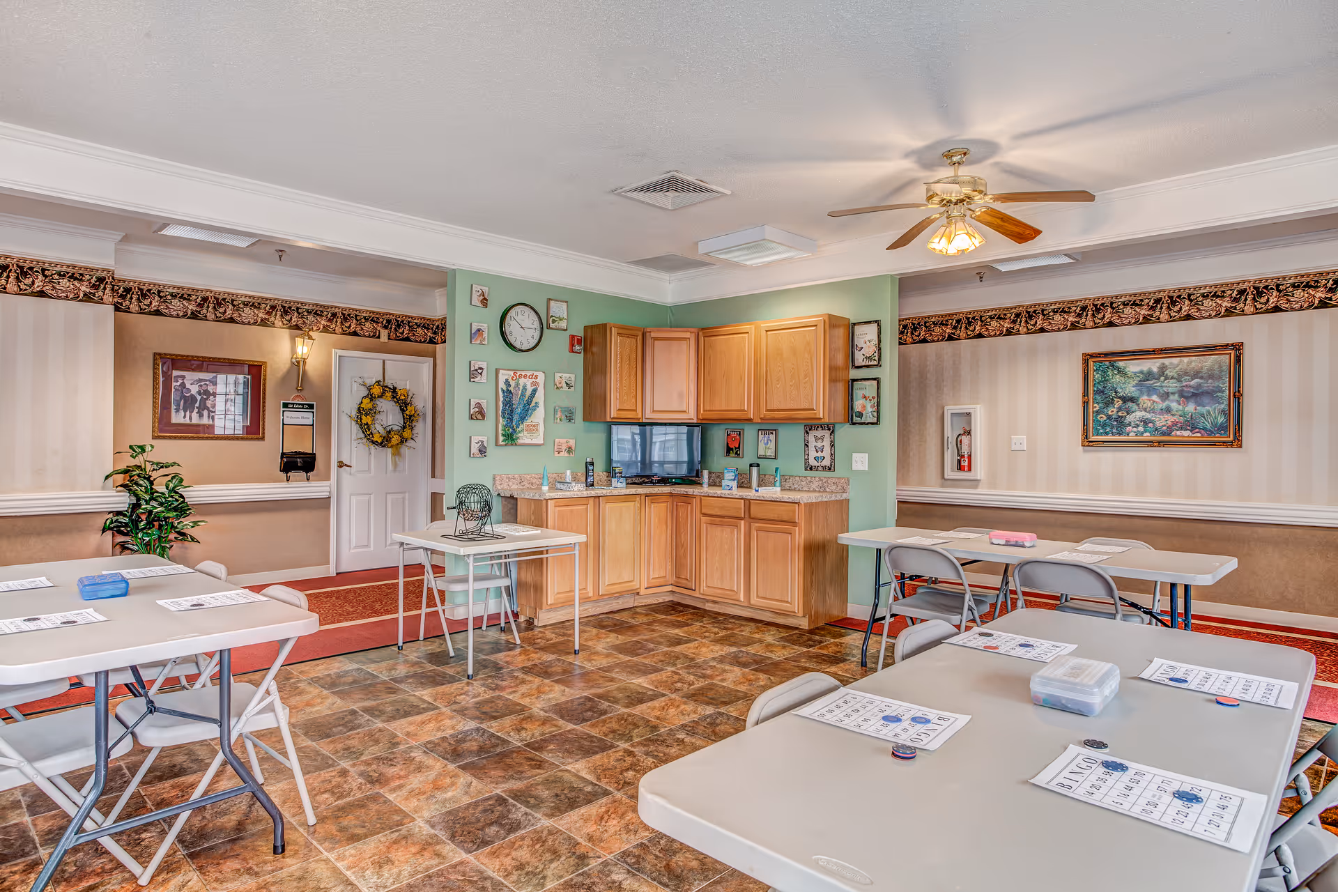 A bright and clean common room in a senior living facility with several folding tables and chairs arranged for activities. Bingo cards and chips are placed on the tables. The room features a small kitchenette with wooden cabinets, a clock, and decorative wall art. There is a ceiling fan with lights, patterned wallpaper, and a door decorated with a wreath.