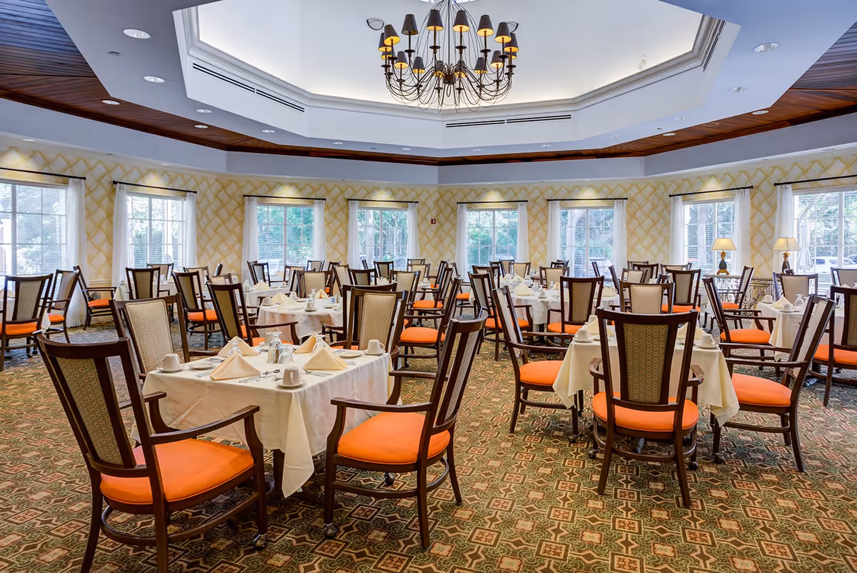 A spacious dining room with multiple round tables covered with white tablecloths, each set with napkins, cups, and silverware. The chairs have wooden frames with orange cushions. Large windows with white curtains allow natural light to fill the room. A decorative chandelier hangs from the ceiling.