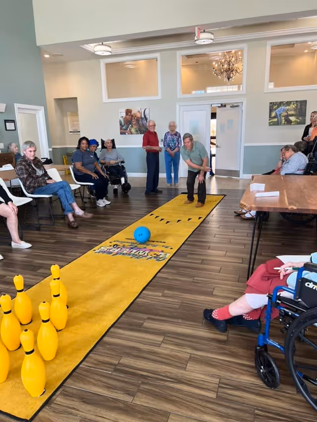 A group of elderly people and caregivers are gathered in a spacious room with wood flooring, watching a woman roll a blue ball down a yellow indoor bowling lane towards yellow bowling pins. Some people are seated in chairs and wheelchairs, and the room has light-colored walls with framed pictures and a chandelier visible through an open doorway.