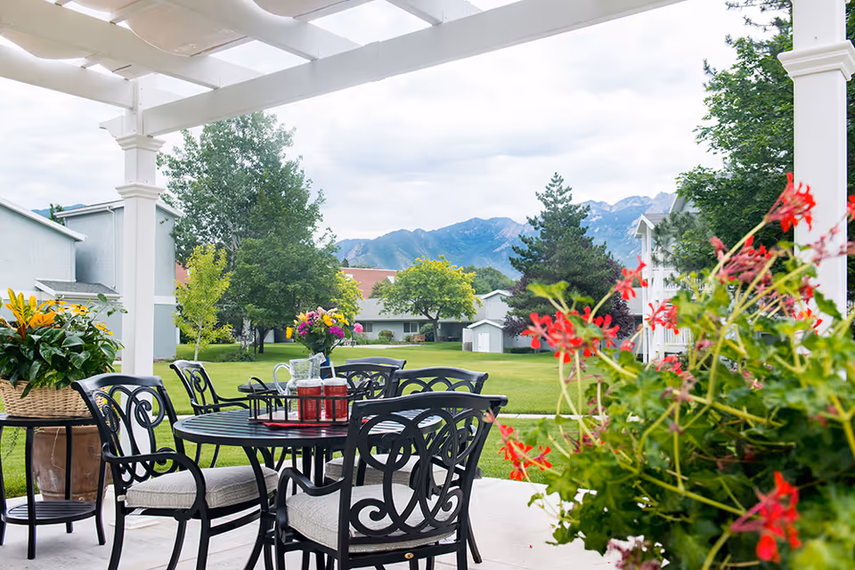 Outdoor patio area with a black metal table and chairs under a white pergola. The table has a tray with a pitcher and glasses, and a vase with colorful flowers. Surrounding the patio are green lawns, trees, and residential buildings with mountains visible in the background under a cloudy sky.