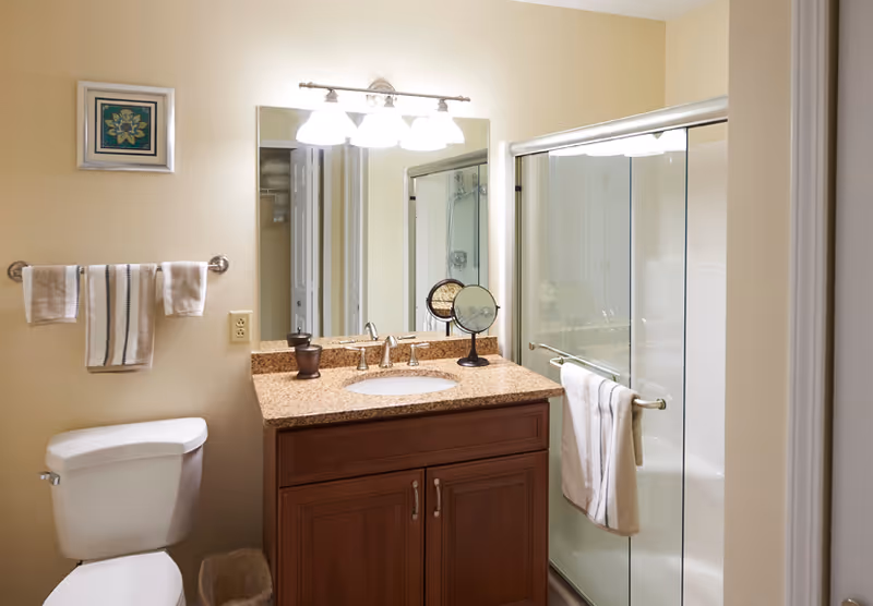 A clean bathroom with a wooden vanity topped with a granite countertop, a sink, and a mirror above it. There is a toilet to the left with a towel rack above holding two towels. To the right is a shower with sliding glass doors and a towel hanging on the handle. The walls are painted beige and there is a small framed artwork on the wall.