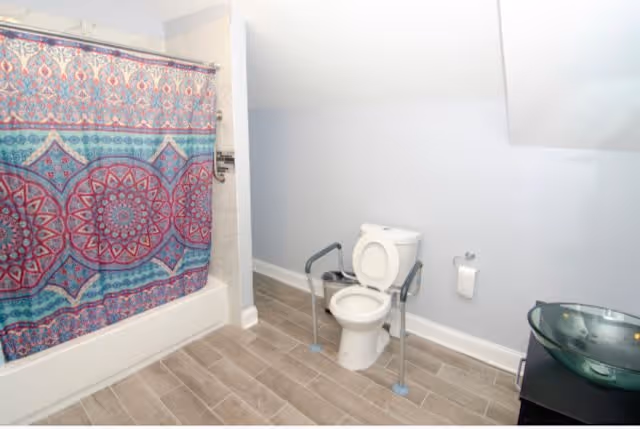 A bathroom with a toilet equipped with safety rails, a colorful patterned shower curtain, a glass vessel sink on a dark vanity, and light gray walls with wood-look tile flooring.