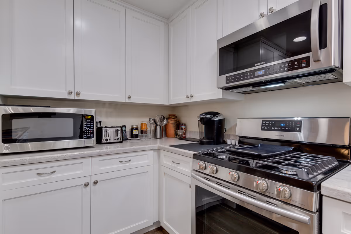 Modern kitchen corner with white cabinets, a stainless steel microwave mounted above a gas stove, a microwave oven on the countertop, a toaster, a coffee maker, and various kitchen utensils and containers.