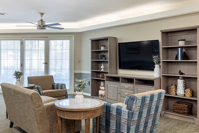 A cozy living room with a ceiling fan, large windows with sheer curtains, a flat-screen TV mounted on a wooden entertainment center, two bookshelves with decorative items, a round wooden coffee table with a white vase, and four upholstered chairs arranged around the table.