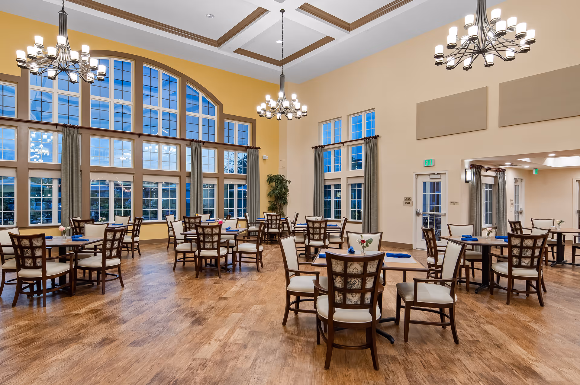 A spacious dining room with multiple wooden tables and chairs arranged neatly. The room features large windows with an arched design letting in natural light, yellow walls, and elegant chandeliers hanging from a coffered ceiling. Blue napkins and small flower arrangements are placed on the tables.