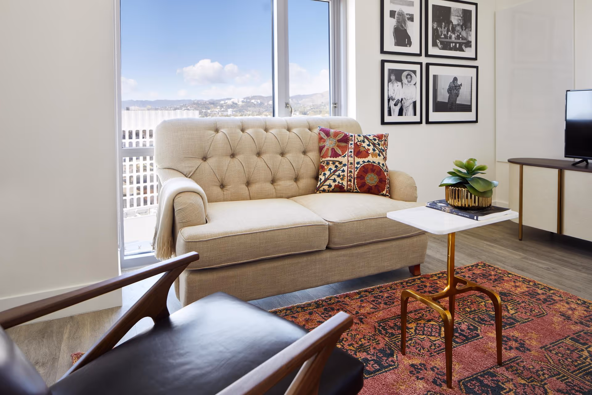 A cozy living room with a beige tufted loveseat adorned with two colorful patterned pillows, a small white marble-top side table with a gold base holding a potted plant and a book, a dark leather armchair with wooden arms, a red patterned rug, a TV on a wooden console, and a wall decorated with four black and white framed photographs. Large windows in the background show a view of buildings and hills under a blue sky with clouds.