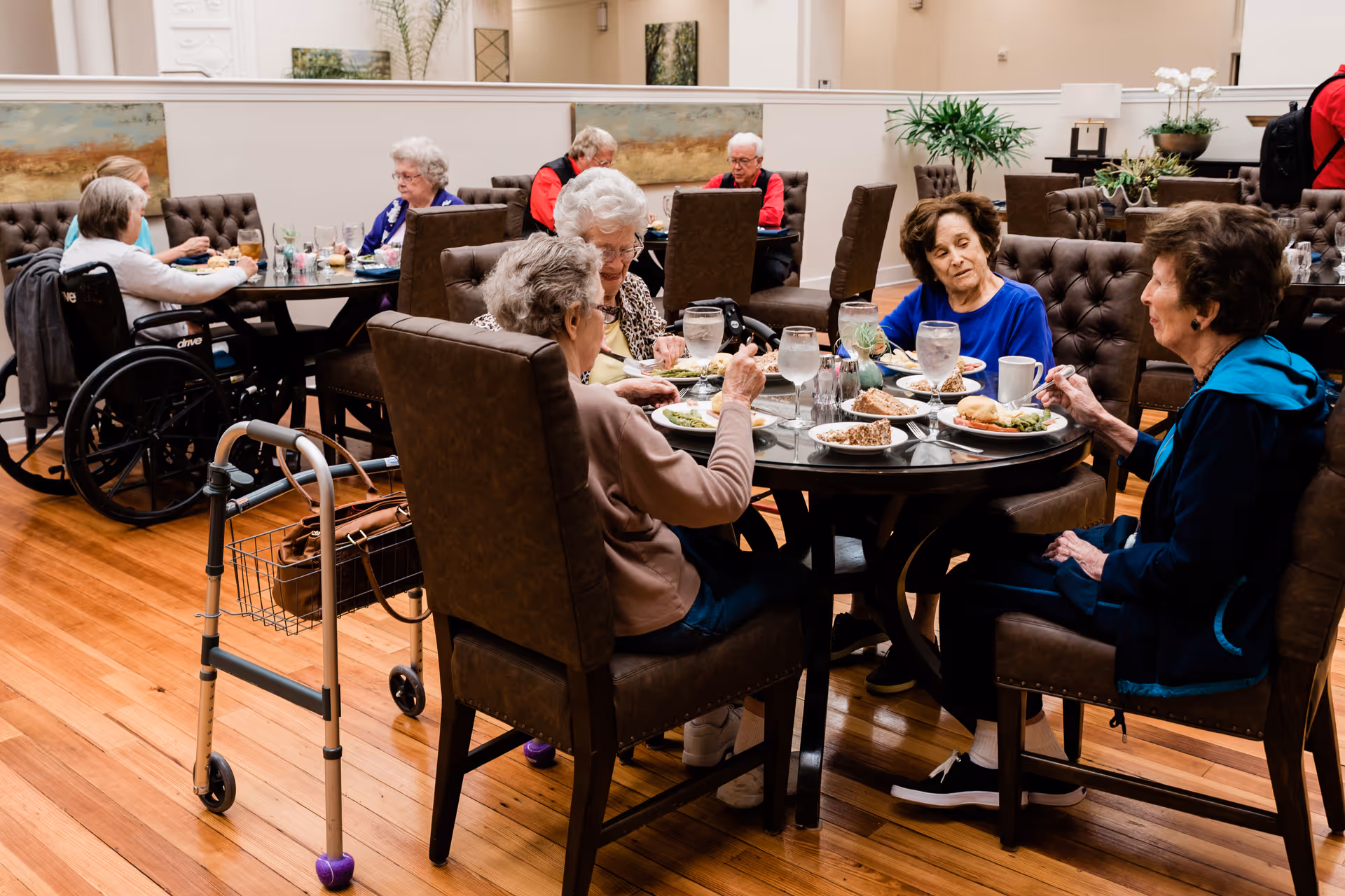 Several elderly women sitting around a round dining table in a senior living facility, eating a meal together. The room has wooden floors, brown cushioned chairs, and a walker is placed near one of the chairs. Other tables with elderly residents are visible in the background.