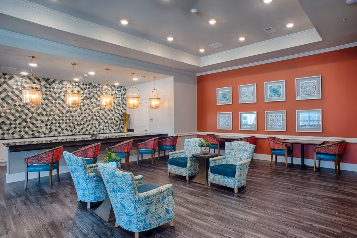 A spacious, well-lit common area with a seating arrangement of four patterned blue armchairs around a small table. Behind the seating area is a long counter with six red and blue chairs, hanging pendant lights, and a decorative tiled wall. The far wall is painted orange and decorated with eight framed artworks arranged in two rows.