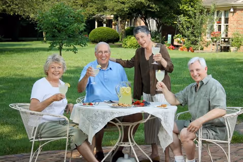 Four elderly adults sitting and standing around a white metal outdoor table with a lace tablecloth, enjoying lemonade and smiling in a green garden area with trees and a brick building in the background.