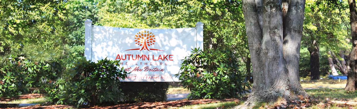 A white sign reading "Autumn Lake Healthcare at New Britain" stands among shrubs and trees on landscaped grounds.