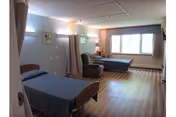 A spacious room with two single beds covered in blue bedspreads, a comfortable armchair between the beds, a window with curtains letting in natural light, and a wooden floor. The room has soft lighting and a calm, clean atmosphere.