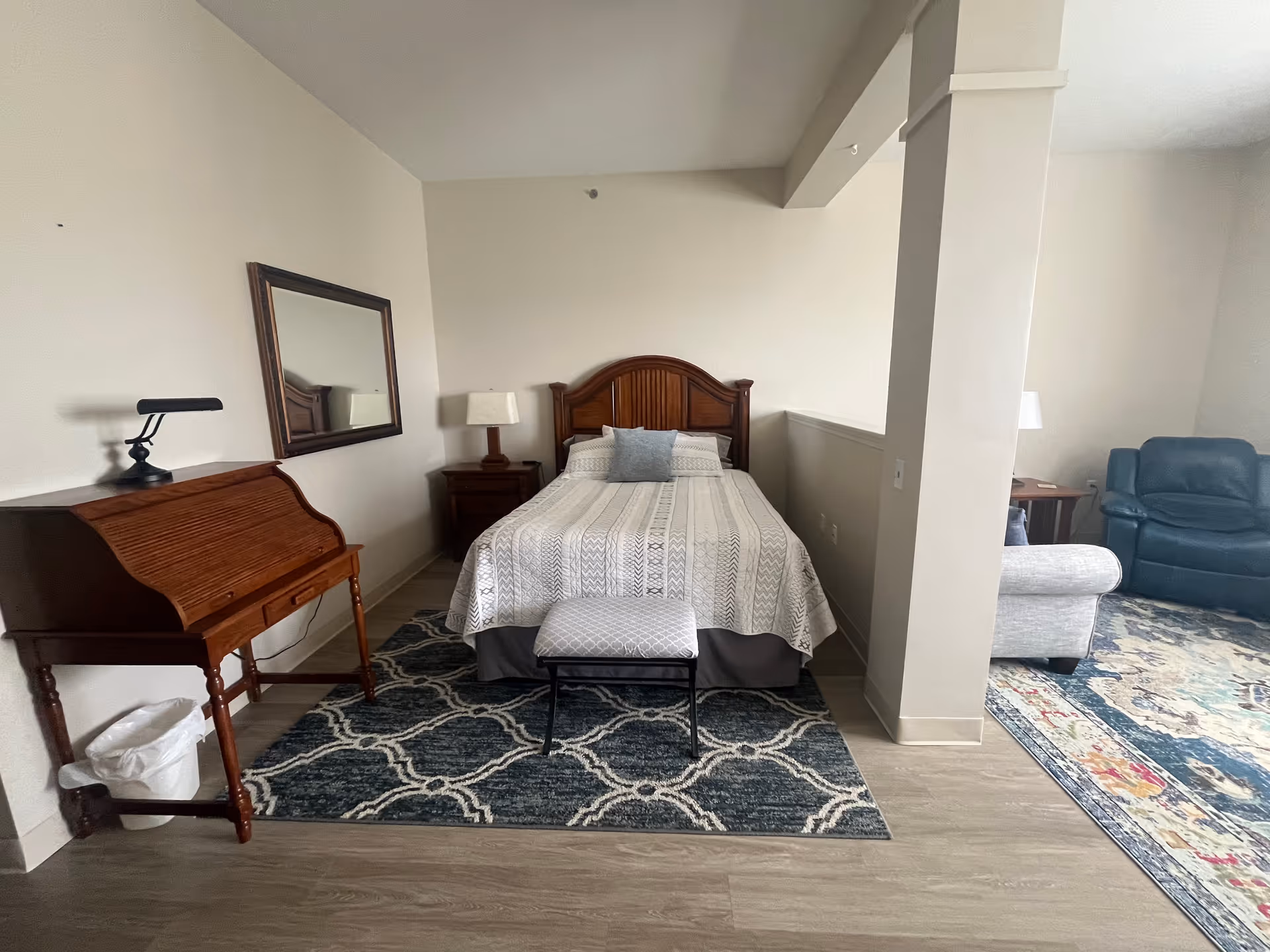 A bedroom area with a wooden bed frame, a bed covered with a patterned quilt and a gray pillow, a wooden nightstand with a lamp, a wooden roll-top desk with a small lamp on it, a mirror on the wall above the desk, and a small upholstered bench at the foot of the bed. Adjacent to the bedroom area is a living space with a gray armchair, a blue recliner, a wooden side table, and a colorful area rug.