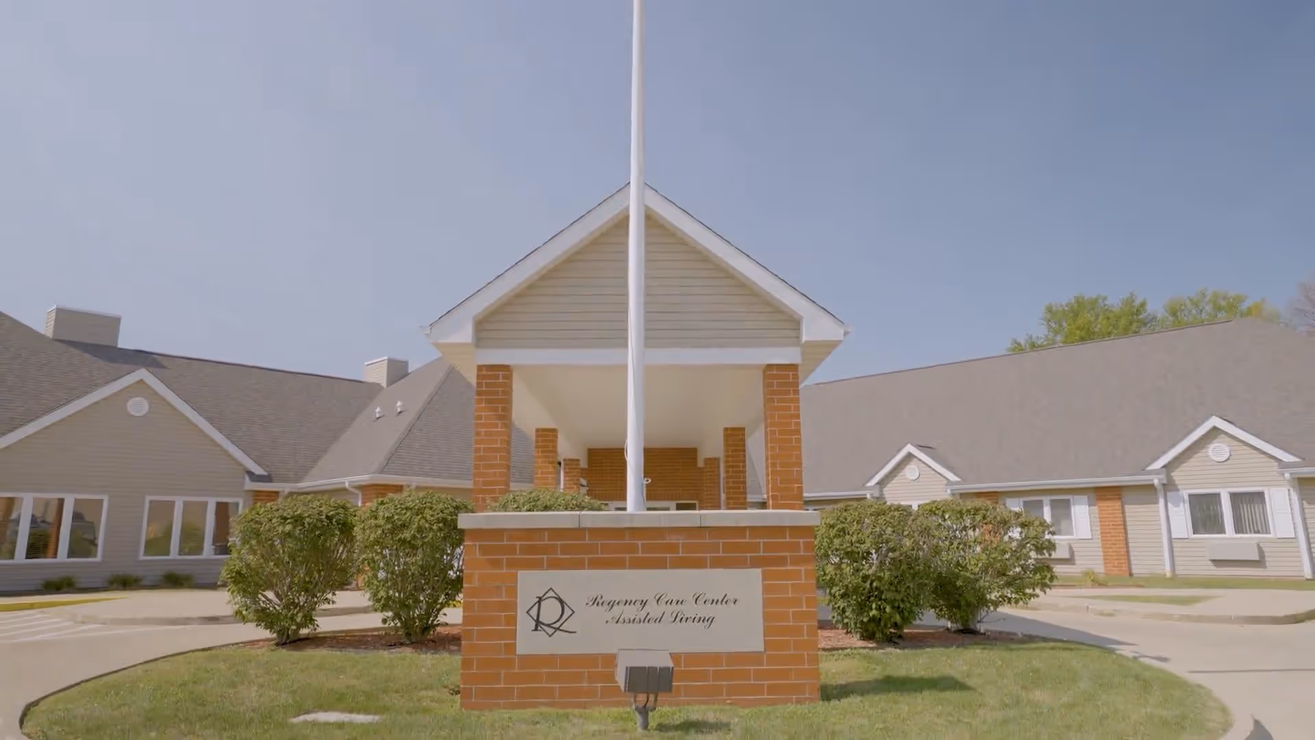 Front exterior view of Regency Care Center Assisted Living building with a brick entrance structure, a flagpole, and neatly trimmed bushes under a clear blue sky.