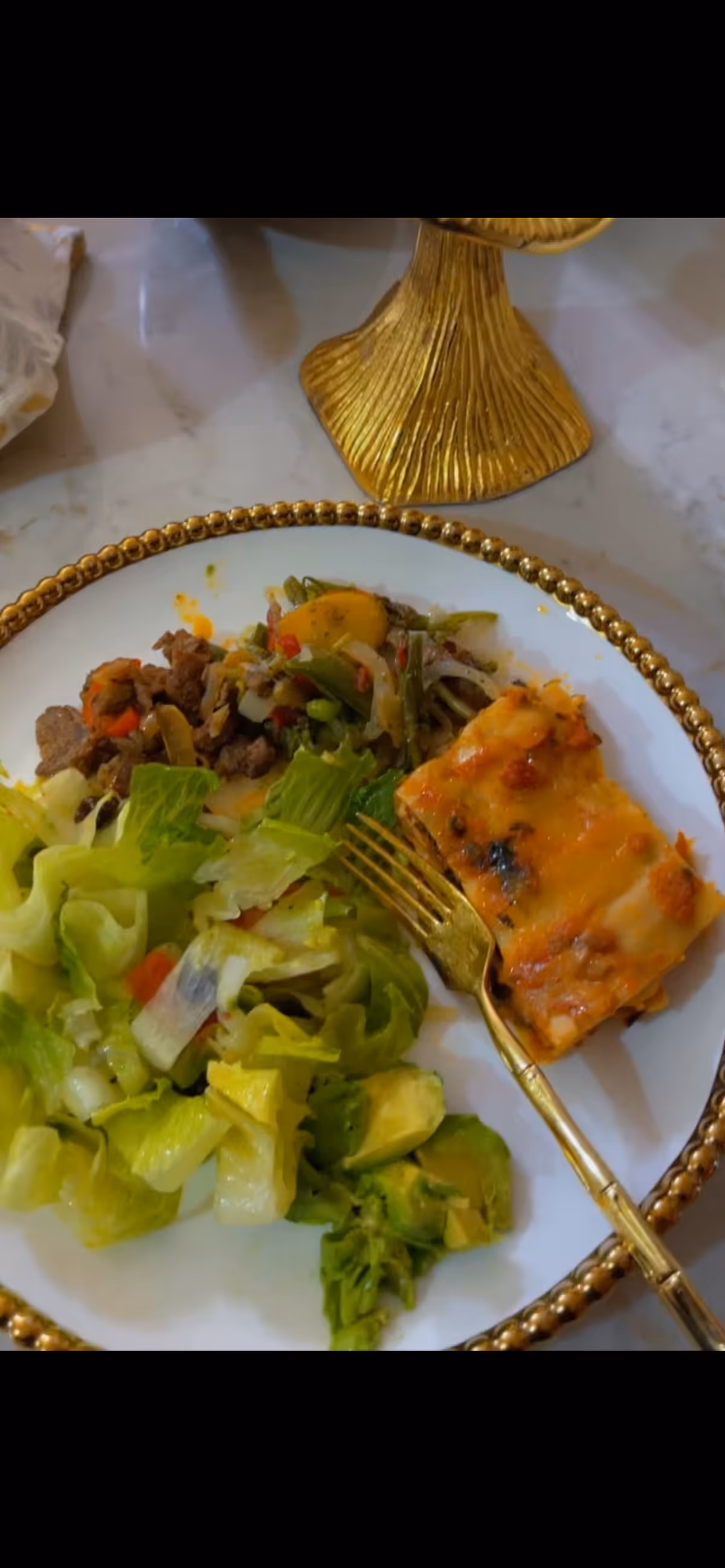 A white plate with a gold beaded rim containing a serving of salad with lettuce and avocado, a portion of cooked mixed vegetables with beef, and a piece of baked lasagna. A gold fork rests on the plate, and a gold decorative object is partially visible in the background.