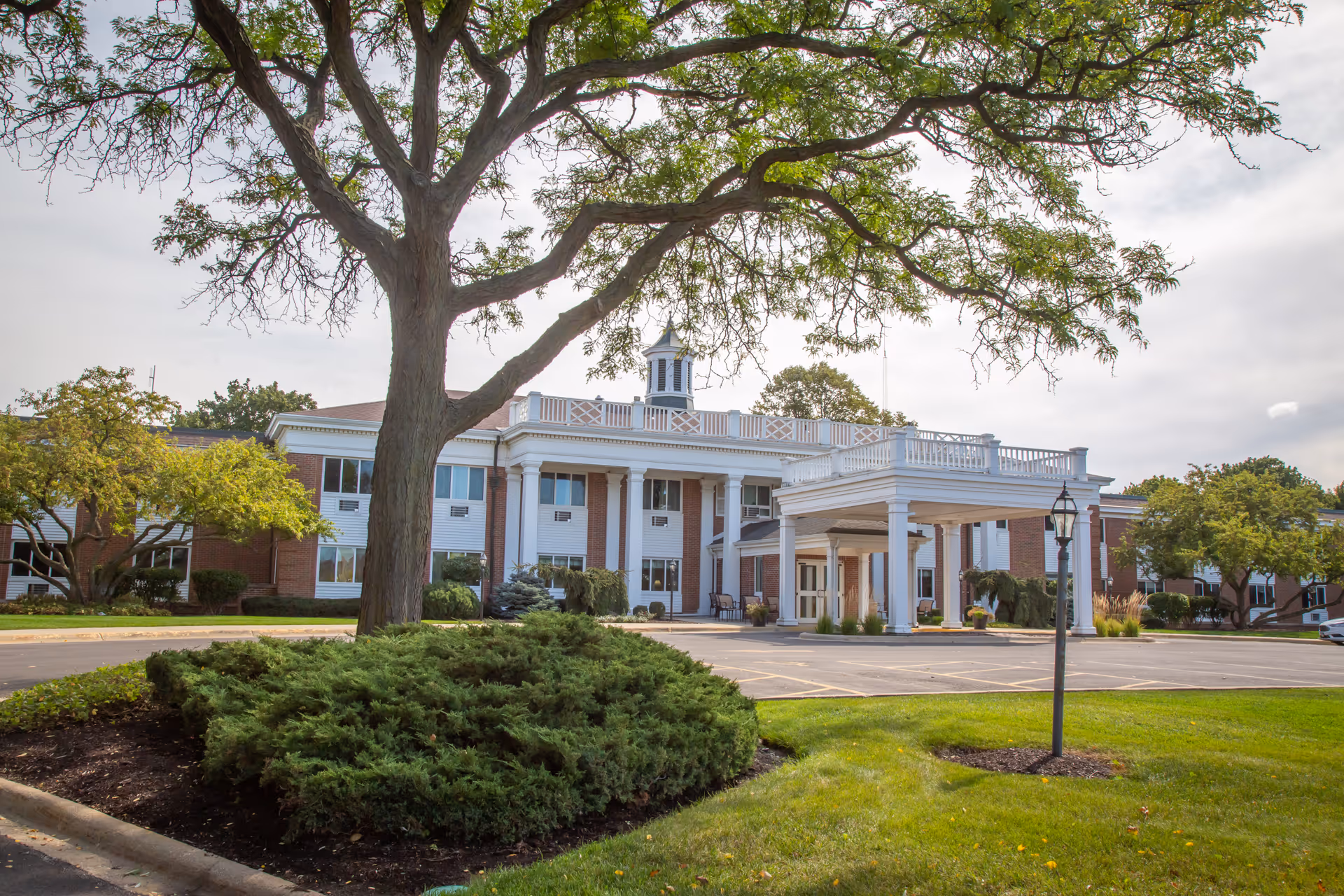 Front entrance of a large brick senior living building with white columns, a covered porte-cochère, and trees and landscaping in the foreground.