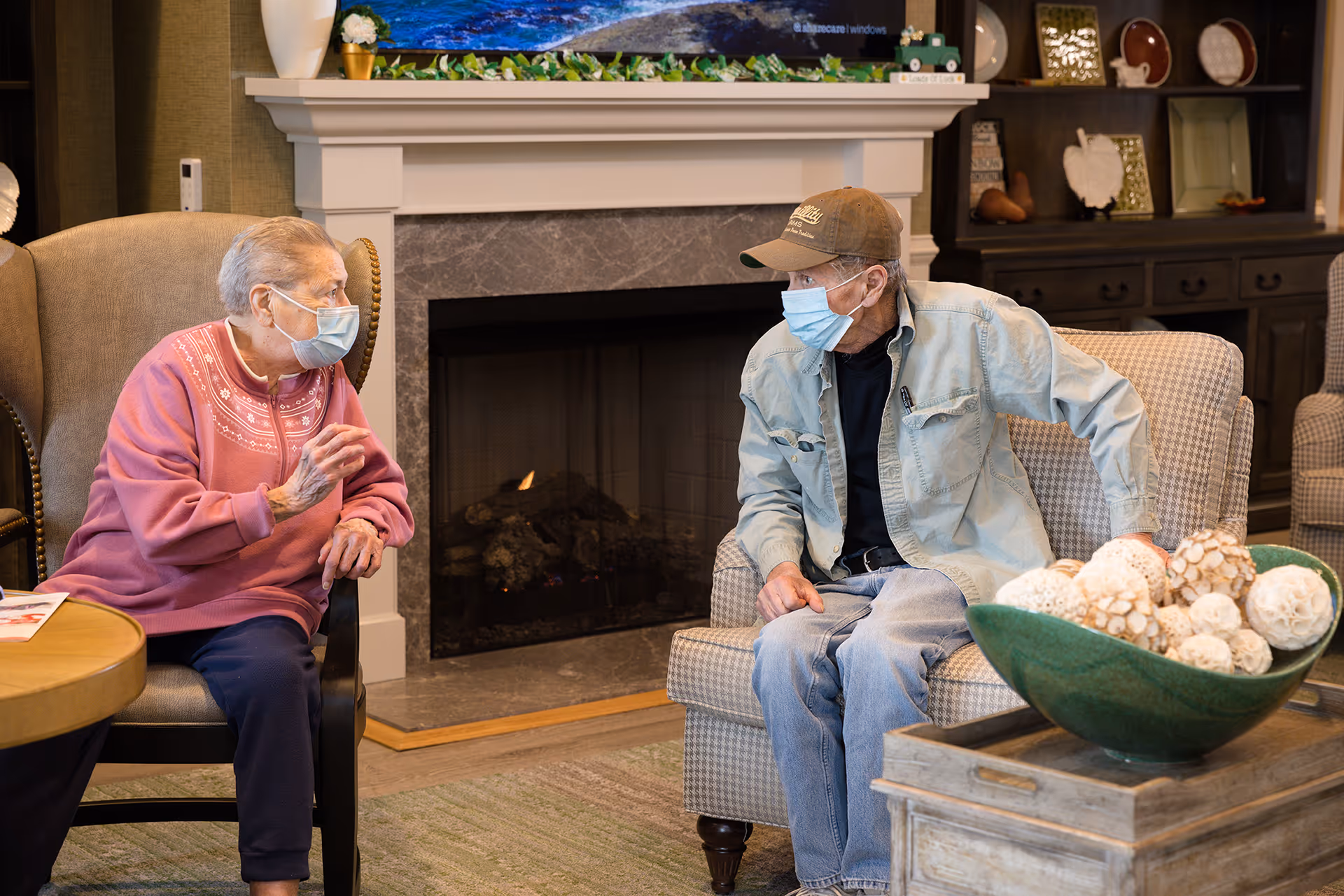 Two elderly people wearing face masks sitting and talking in a cozy living room with a fireplace in the background. One person is seated in a cushioned armchair wearing a pink sweater, and the other is seated on a patterned chair wearing a cap and denim jacket. A decorative bowl with white spherical objects is on a wooden table in the foreground.