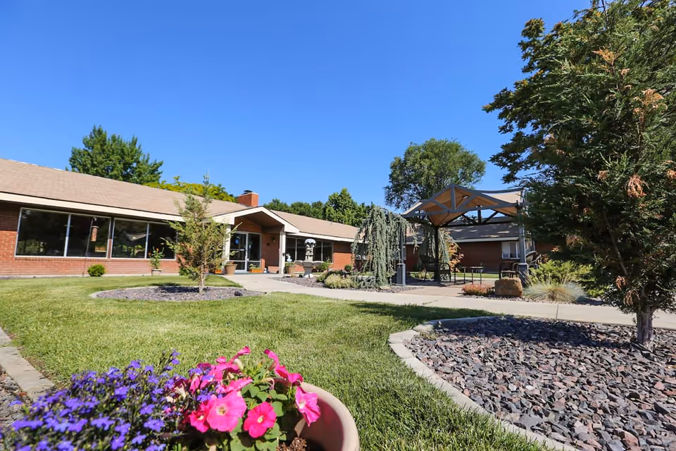 Outdoor view of Heritage Assisted Living of Boise showing a single-story brick building with large windows, a well-maintained lawn, flower beds with pink and purple flowers, trees, and a covered seating area under a pergola on a sunny day with clear blue sky.