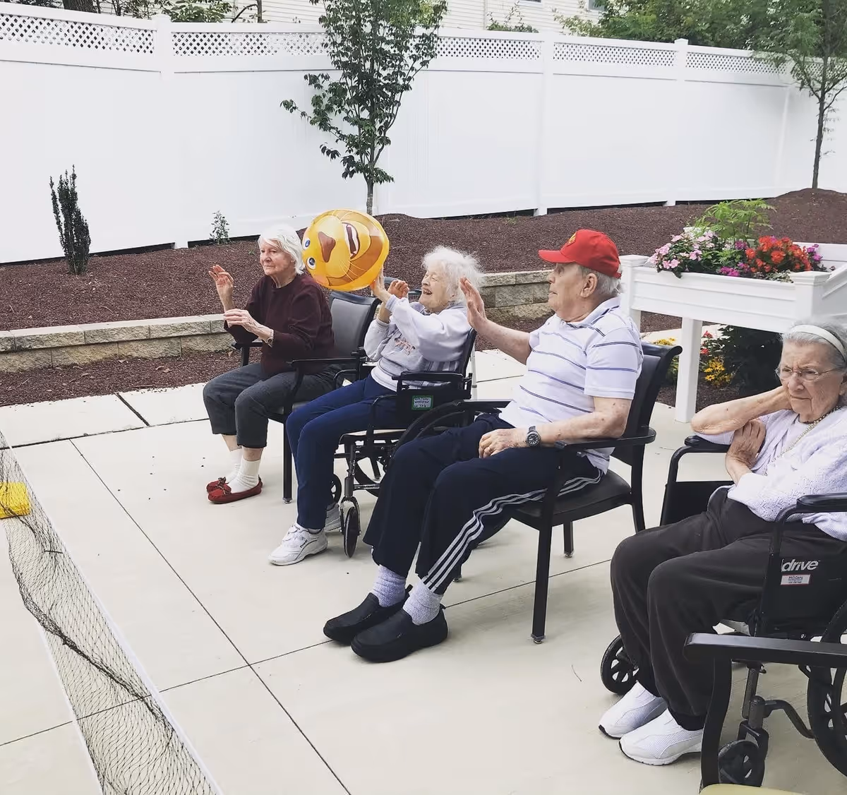 Four elderly individuals sitting outdoors on chairs and wheelchairs, engaging in a light activity with a yellow inflatable ball. They are seated on a paved area with a white fence and some small trees and plants in the background.