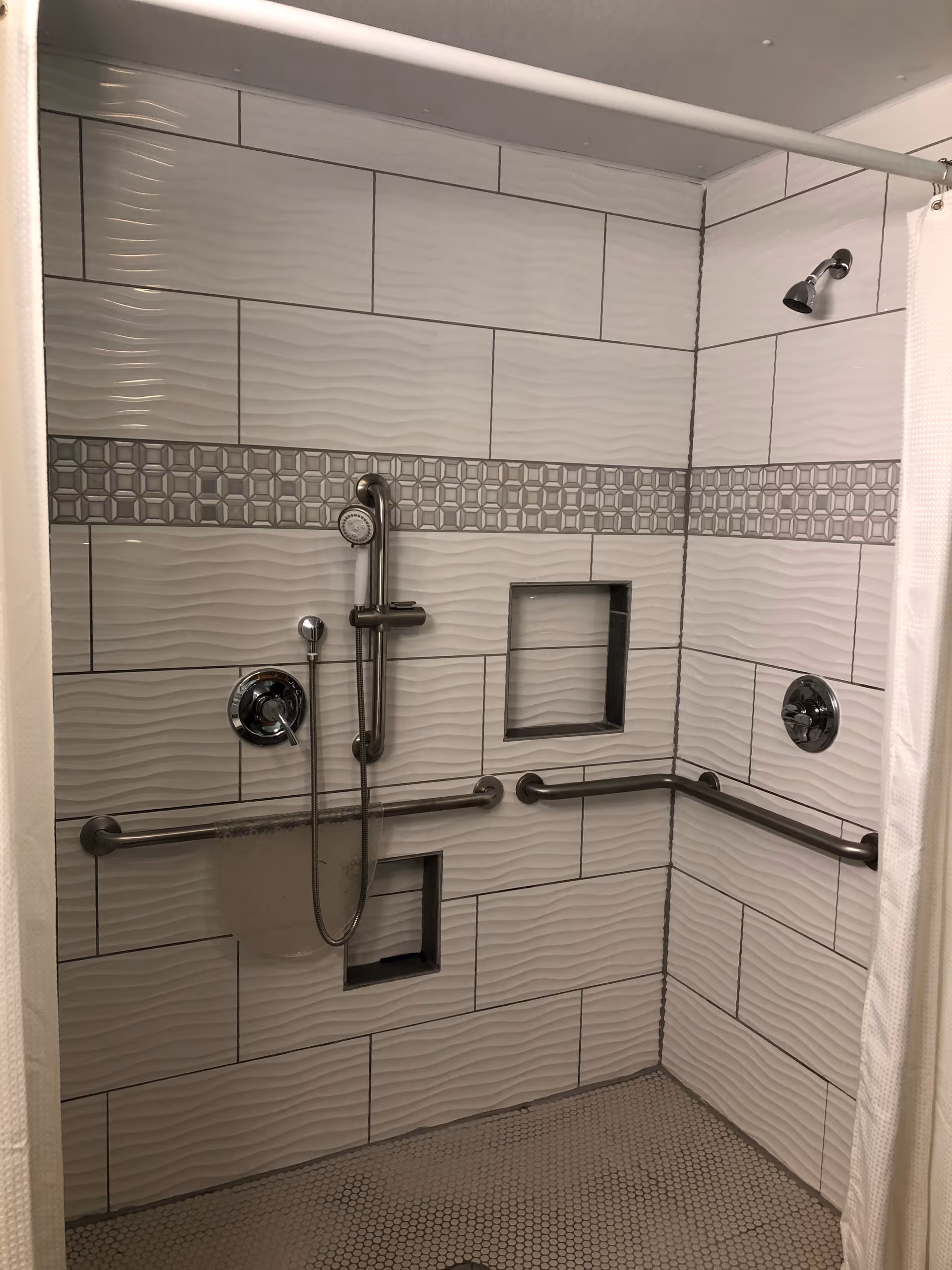 A tiled shower area with white and gray wave-patterned tiles and a decorative horizontal strip of geometric tiles. The shower has two showerheads, one fixed and one handheld attached to a vertical bar. There are two built-in recessed shelves and two horizontal grab bars for accessibility. The floor is covered with small hexagonal tiles, and a white shower curtain is partially visible on the right side.