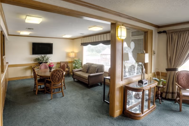 A cozy living room area in a senior living facility with a round wooden table surrounded by four chairs, a brown sofa with cushions, a flat-screen TV mounted on the wall, a floor lamp, and large windows with curtains letting in natural light. There is a wooden display cabinet with decorative items and a small plant on a side table near the window.