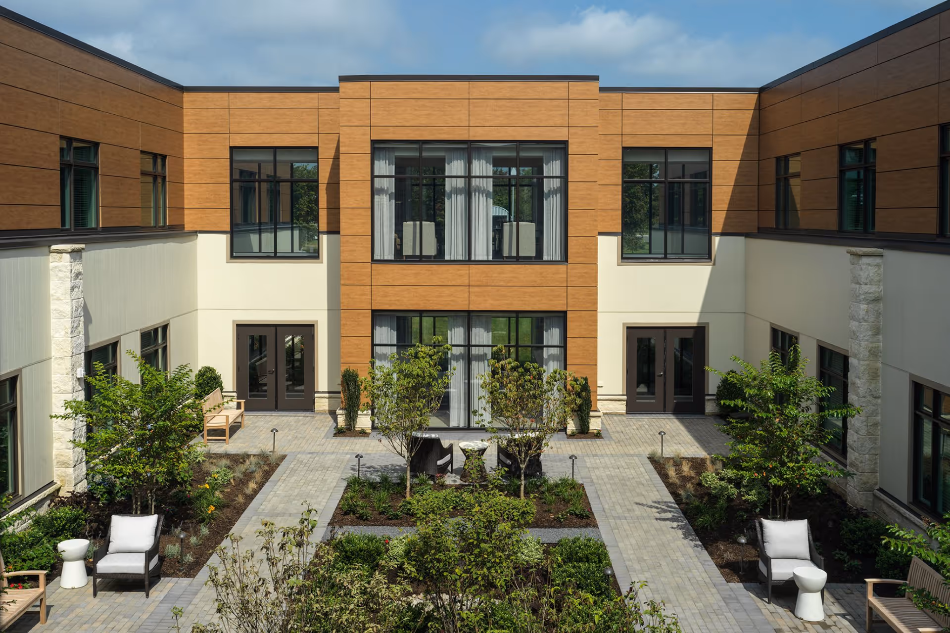 Outdoor courtyard area of a senior living facility with paved walkways, green plants, small trees, benches, and cushioned chairs arranged around small tables. The courtyard is surrounded by a two-story building with large windows and wooden paneling on the upper level.