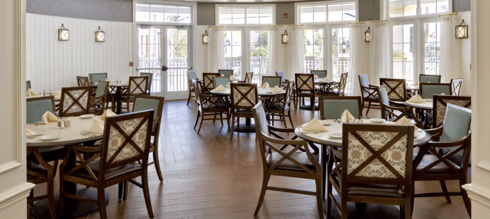 Bright dining room with multiple round tables set with napkins and wooden chairs arranged near large windows and glass doors.