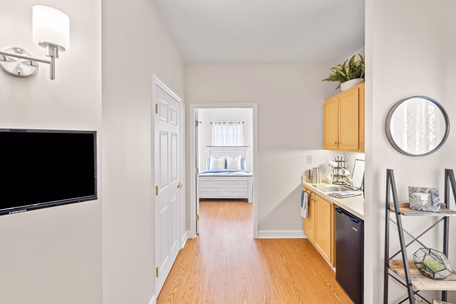 Interior view of a senior living facility unit showing a small kitchenette with wooden cabinets, a mini refrigerator, a sink, and a countertop. To the left, there is a wall-mounted flat-screen TV and a closed white door. Straight ahead, an open doorway leads to a bedroom with a bed featuring white and blue bedding and a window with sheer curtains. On the right side, there is a decorative shelving unit with plants and picture frames, and a round mirror hanging on the wall.