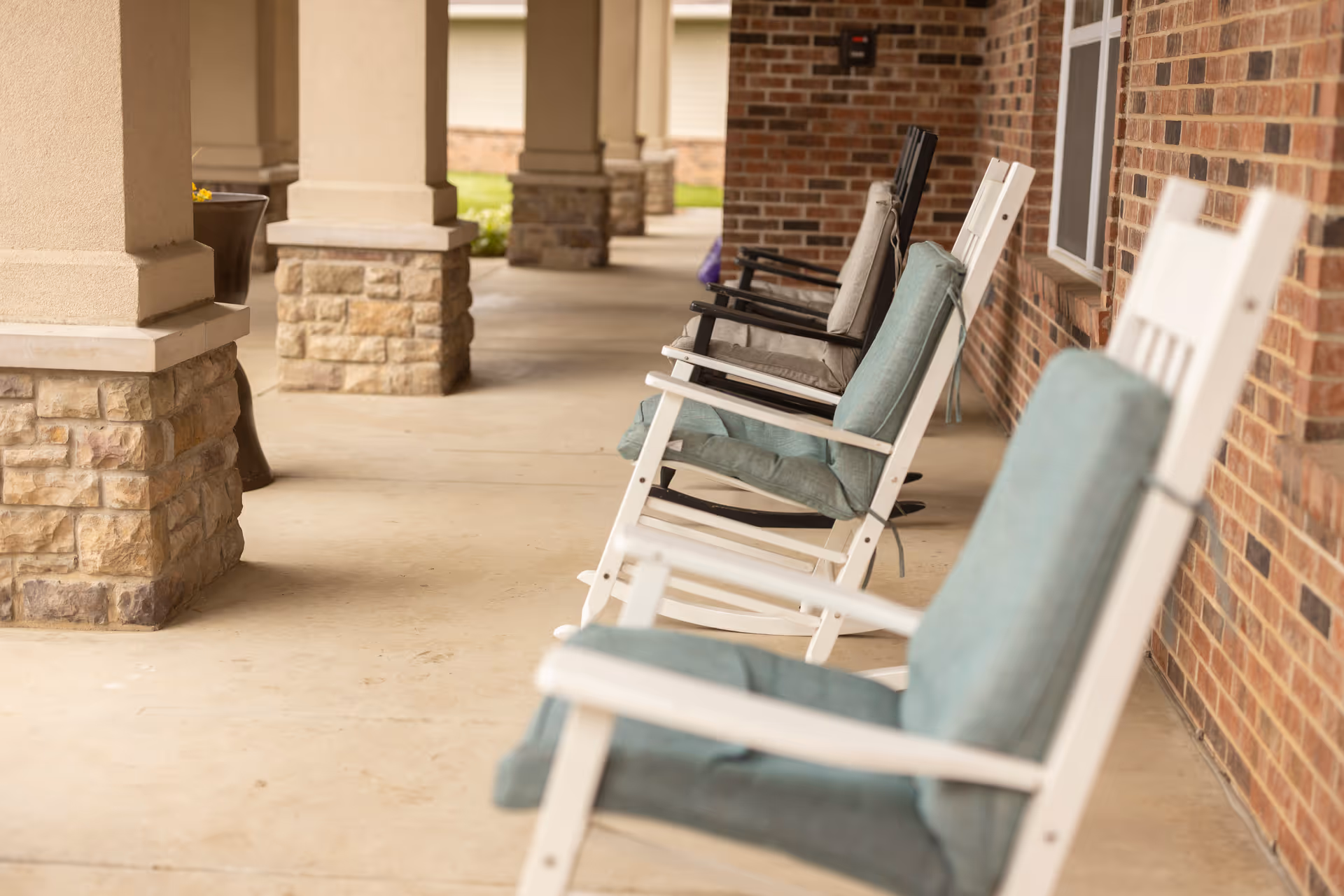 A row of cushioned rocking chairs lined up on a covered porch with stone pillars and a brick wall, overlooking a garden area.