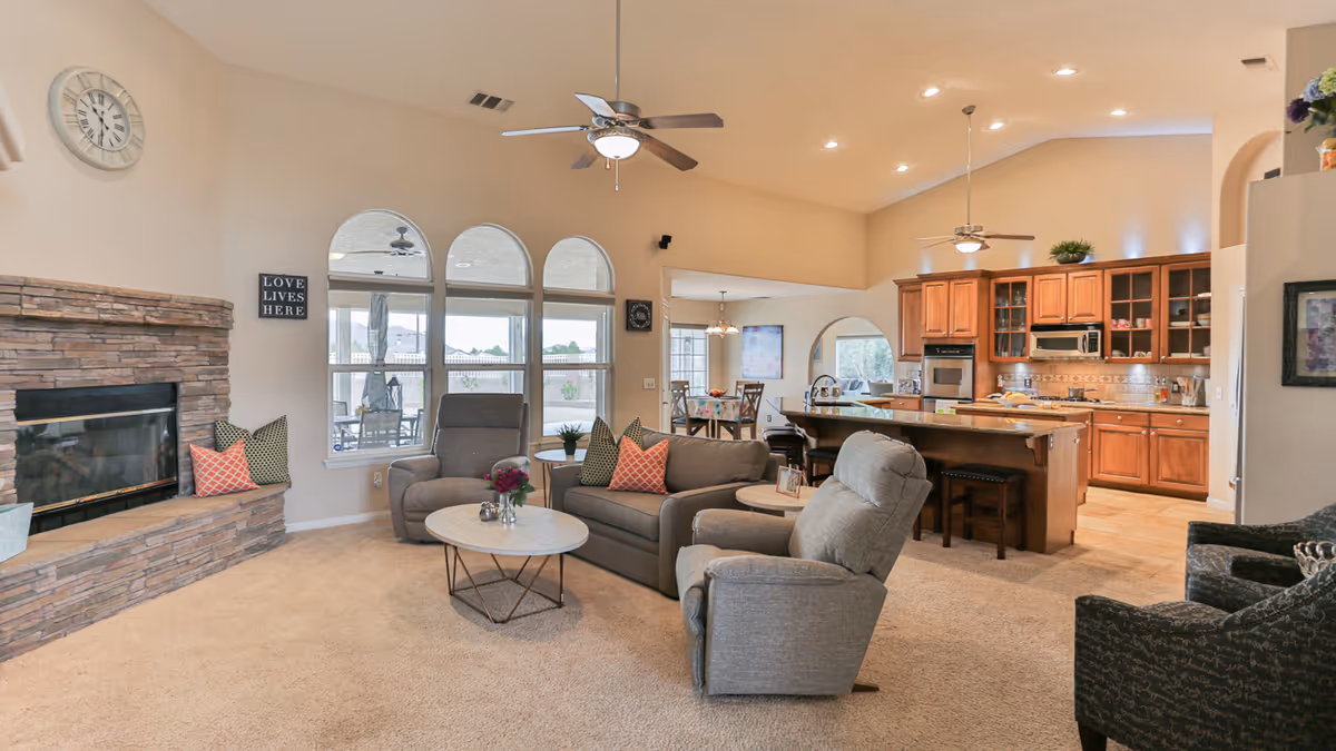 A spacious and well-lit living room area with beige carpet, a stone fireplace on the left, and a seating arrangement including a sofa, two armchairs, and a round coffee table with flowers. The room has large arched windows letting in natural light, ceiling fans, and an open view into a kitchen with wooden cabinets and a dining area in the background.