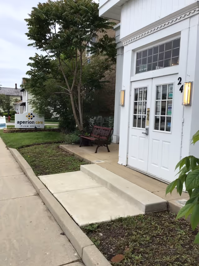 Front entrance of a white building with double doors, a short concrete ramp and bench, a tree, and a sign reading "aperion care Plum Grove" by the sidewalk.