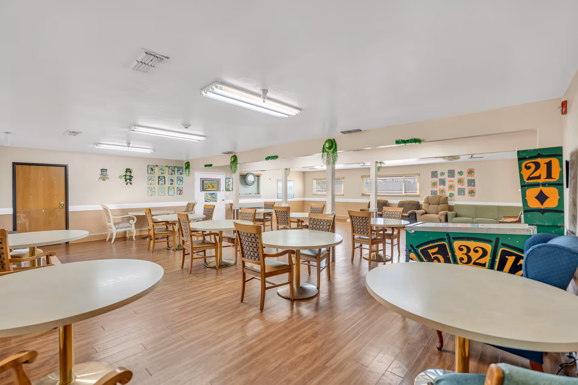 A spacious common area with multiple round tables and chairs arranged on a wooden floor. The room has beige walls decorated with colorful artwork and green hanging decorations. There are several windows letting in natural light, and a few comfortable armchairs and sofas are visible in the background. The ceiling has fluorescent lighting fixtures.