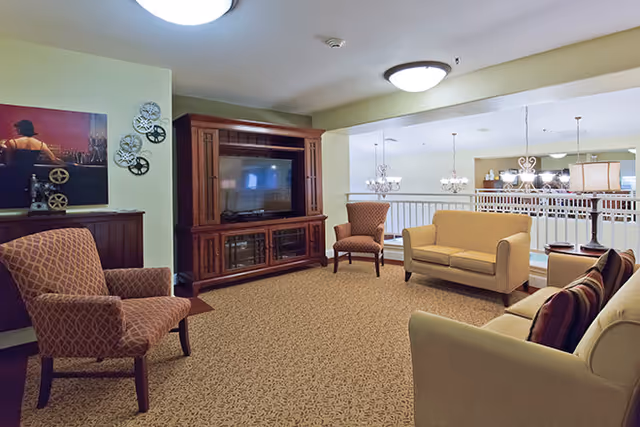 A cozy living room area in a senior living facility with patterned carpet, a wooden entertainment center with a TV, two armchairs, and two sofas. The room is softly lit with ceiling lights and overlooks a dining area with chandeliers.