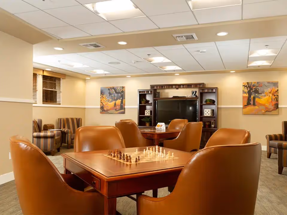 Communal lounge with leather chairs around a table set up for chess, a TV entertainment center, and framed artwork on the walls.