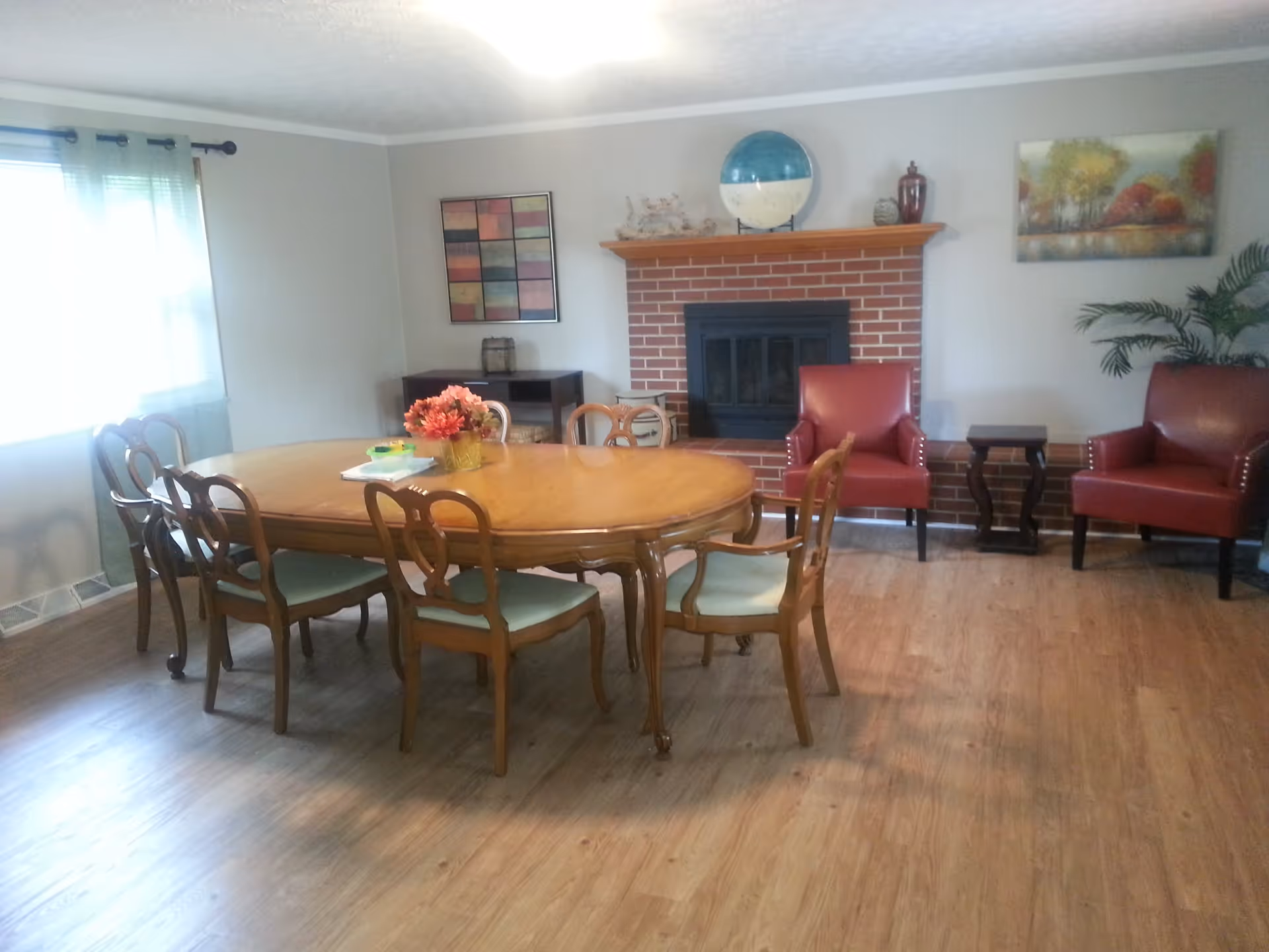 A cozy dining room with a wooden oval dining table surrounded by six chairs with green cushions. On the table is a vase with orange flowers and some papers. The room has a wooden floor, a window with sheer curtains on the left, and a brick fireplace with a wooden mantle on the back wall. The mantle holds a globe, decorative items, and there are two red armchairs with a small wooden side table between them to the right of the fireplace. The walls are decorated with two paintings and a potted plant is visible in the corner.