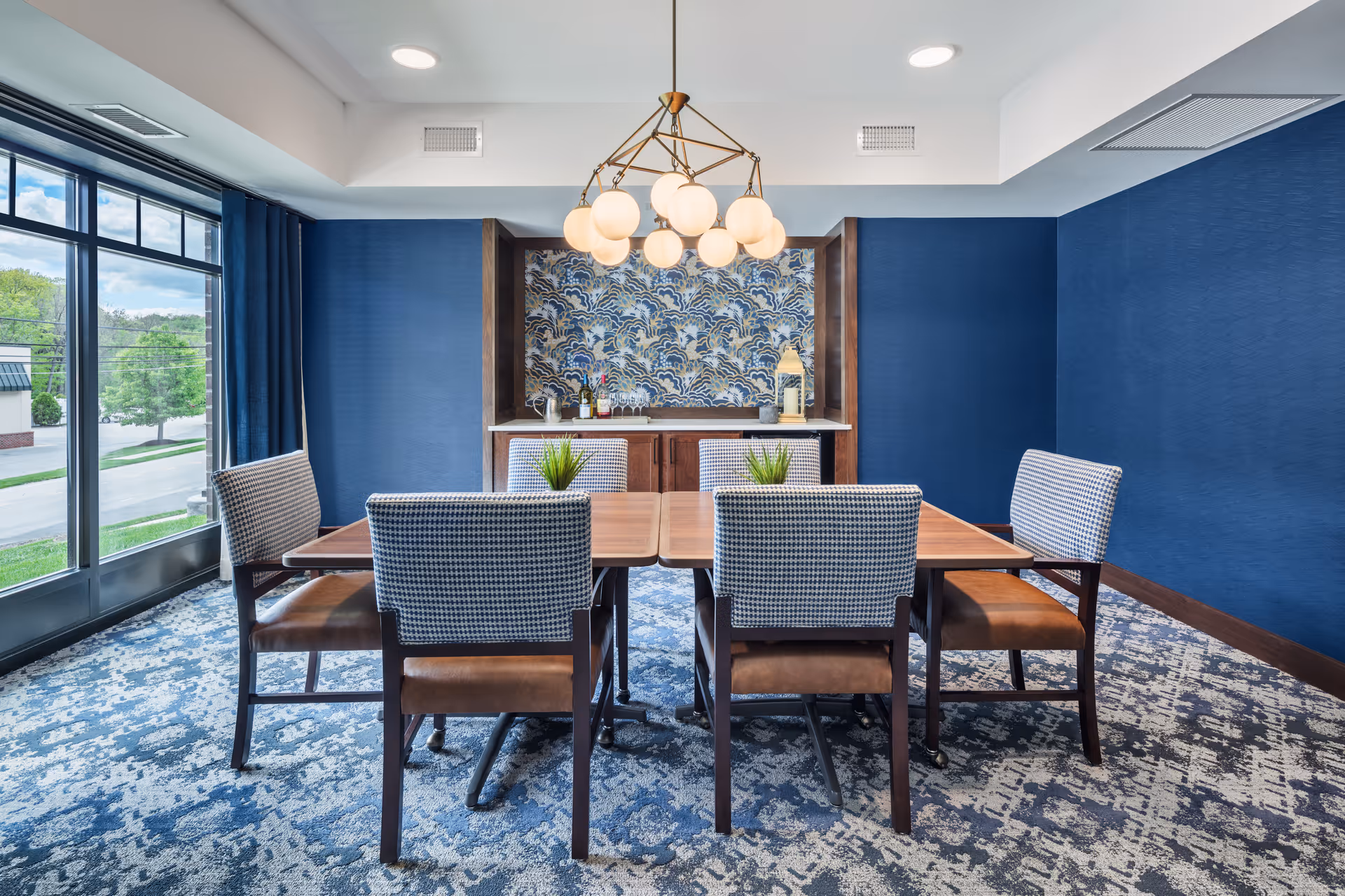 A dining room with a wooden table and six chairs with patterned upholstery and brown seats. The room has blue walls, a patterned carpet, and a large window with blue curtains letting in natural light. A modern chandelier with round white lights hangs above the table. At the back of the room, there is a built-in cabinet with a decorative blue and white patterned backsplash and some bottles and decor items on the countertop.