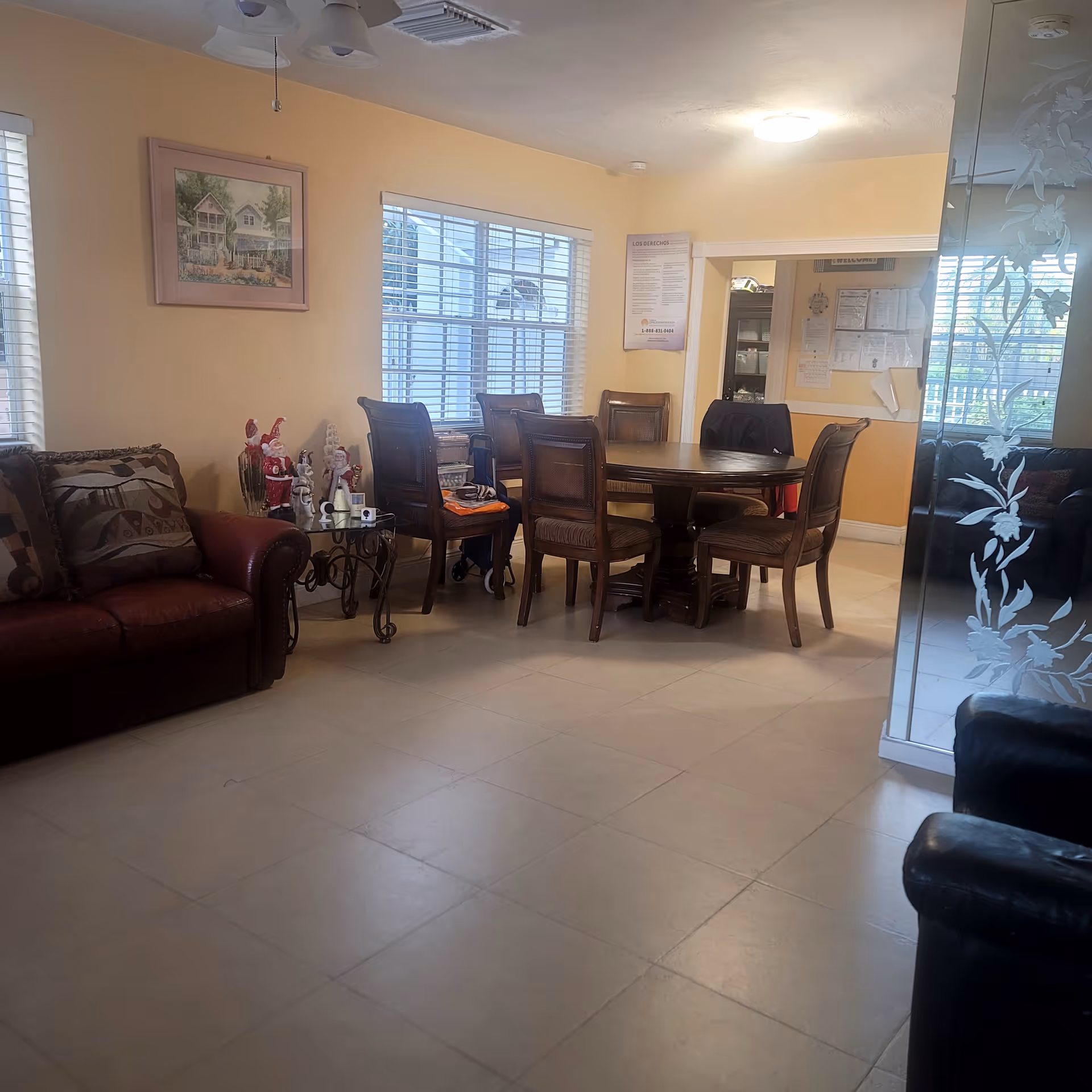 Bright common room with a round dining table and chairs, sofas, and large windows in a senior living facility.