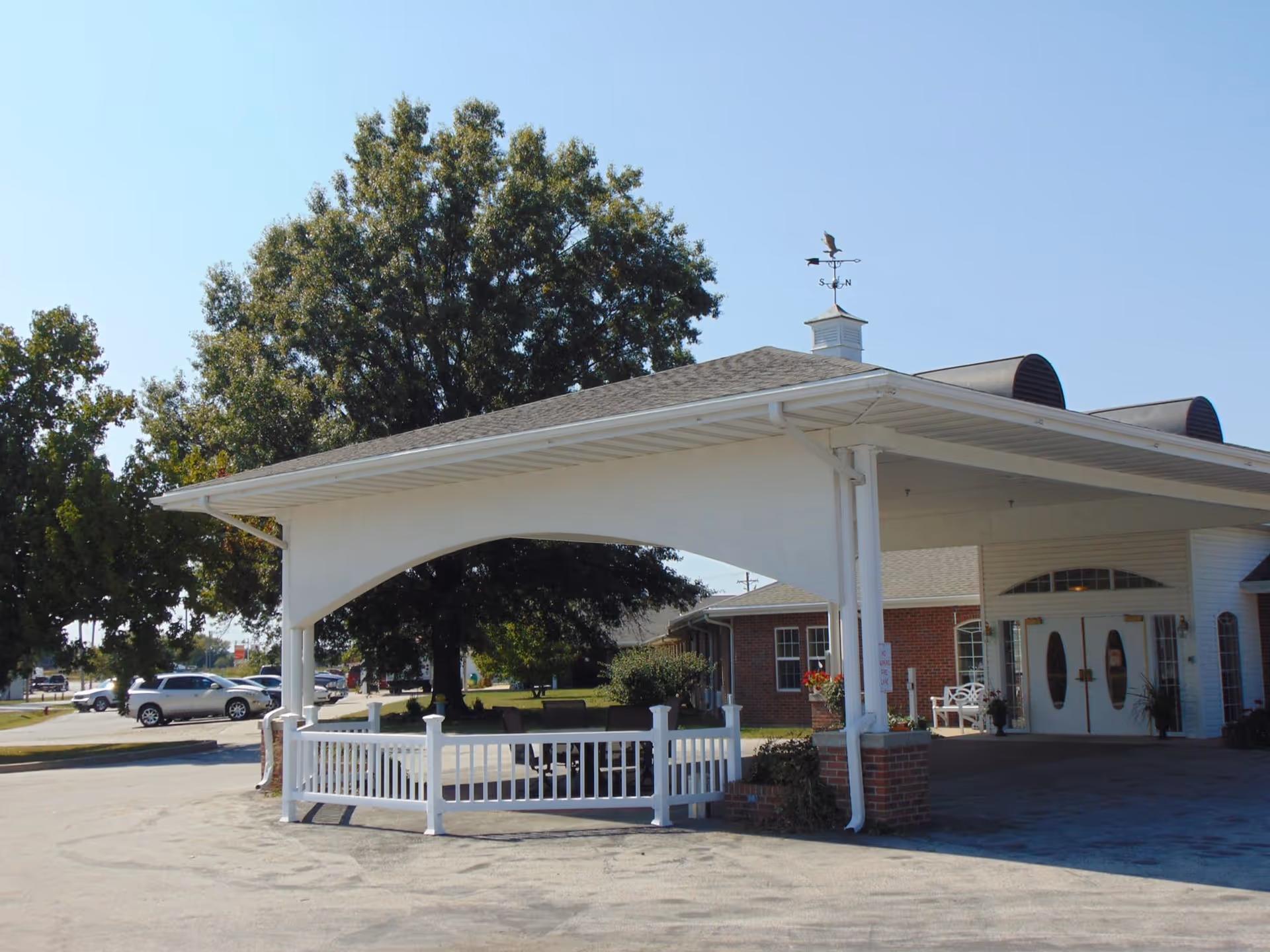 Exterior view of Lincoln Community Care Center & Lakeside Suites showing a covered entrance with white pillars and a small white fence. There are trees and parked cars in the background under a clear blue sky.