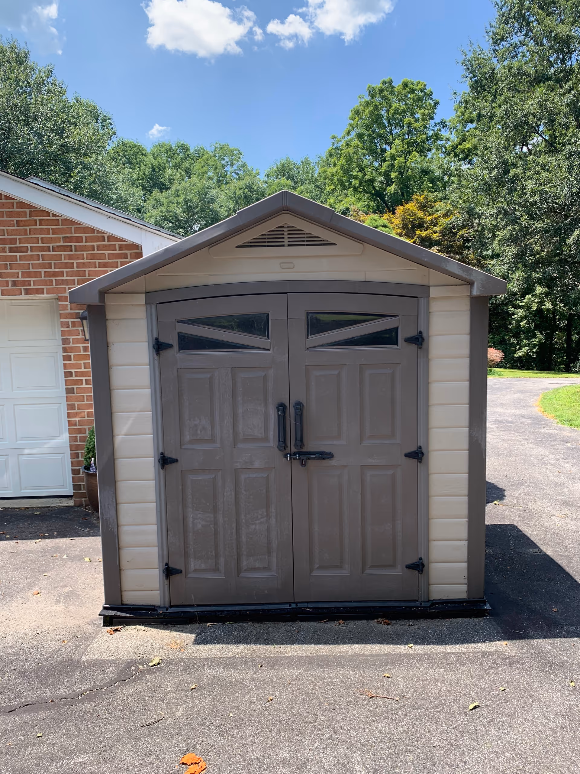 A small brown and beige storage shed with double doors on a driveway next to a brick garage and trees in the background.