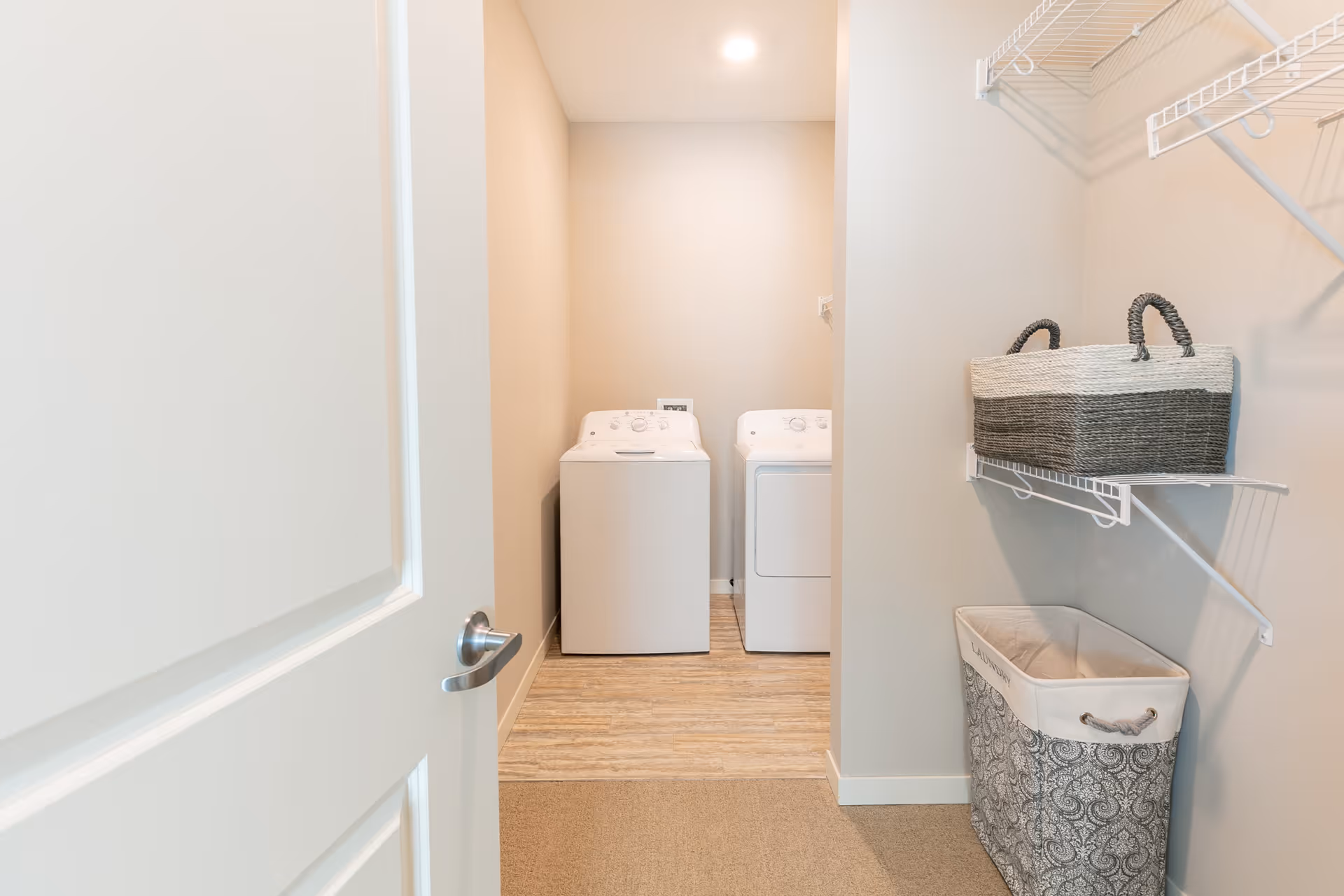 Open doorway revealing a small laundry room with a washer and dryer and wall shelving holding laundry baskets.