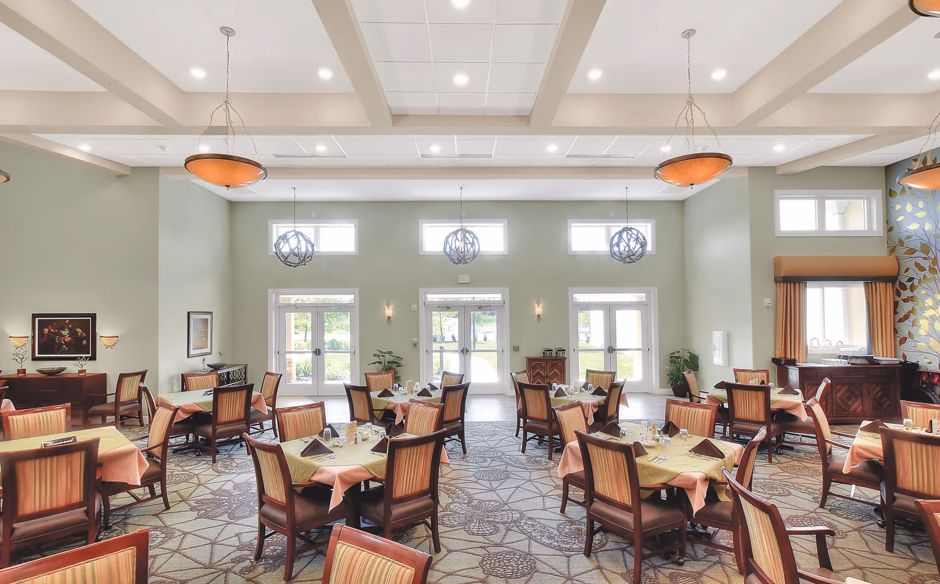 A bright and spacious dining room with multiple tables covered with yellow and peach tablecloths, each set with napkins and silverware. The room features large windows and glass doors letting in natural light, green walls, decorative hanging light fixtures, and patterned carpet. There are wooden chairs with striped cushions around the tables and some framed artwork on the walls.