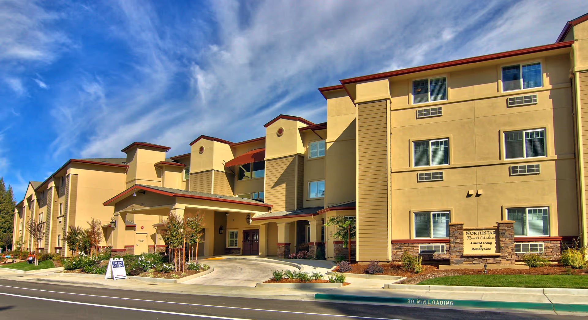 Exterior view of a three-story senior living facility building with beige walls and red trim under a blue sky with wispy clouds. The entrance features a covered drop-off area with a driveway and landscaped plants and trees around the building. A sign near the entrance reads 'Northstar Rancho Cordova Assisted Living & Memory Care.'