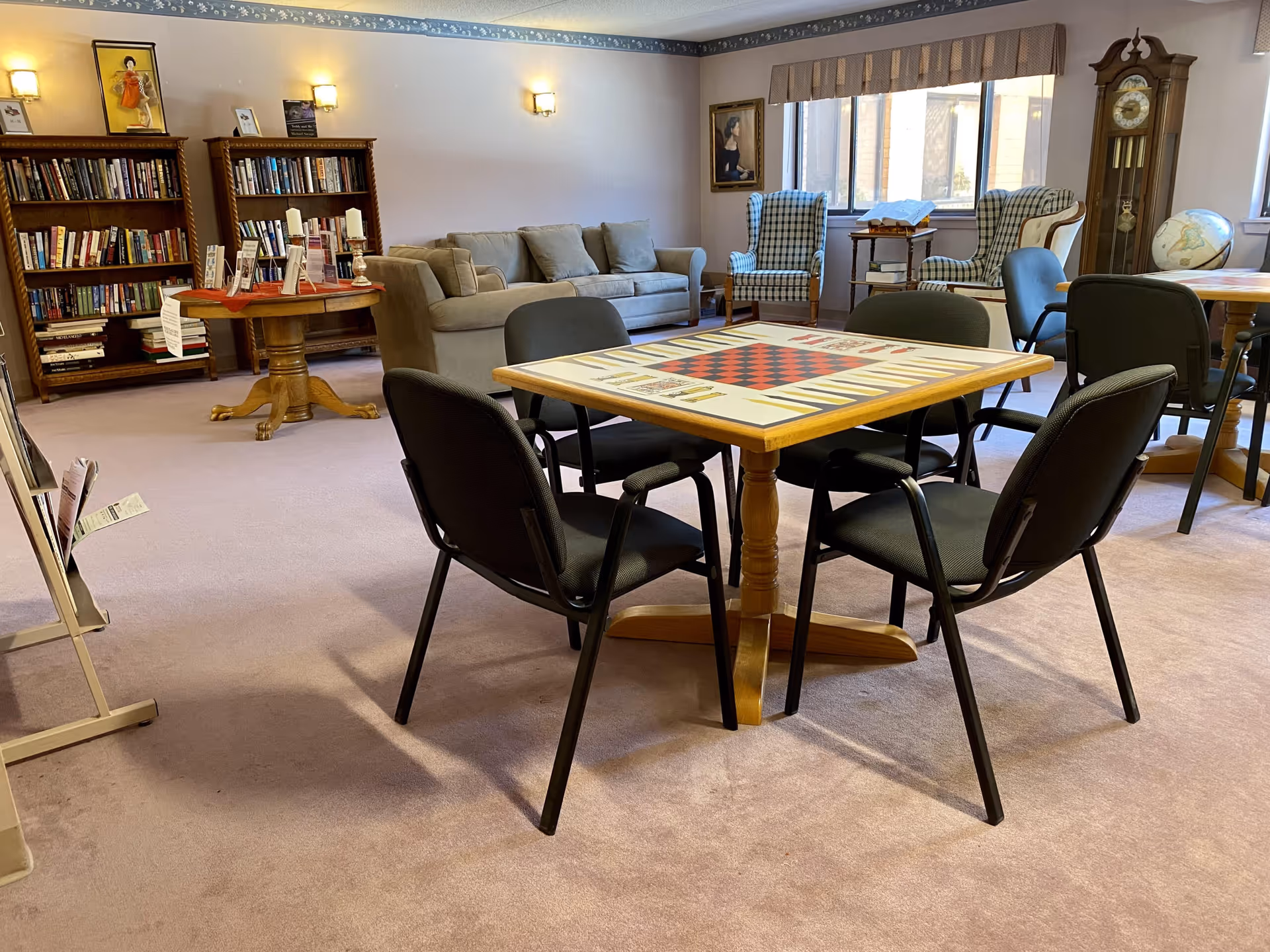 A cozy common area in a retirement community featuring a square game table with a checkerboard and backgammon design surrounded by four black chairs. In the background, there are two wooden bookshelves filled with books, a beige sofa, two checkered armchairs near a window, a grandfather clock, a globe, and framed artwork on the walls. The room has soft lighting and carpeted floors.
