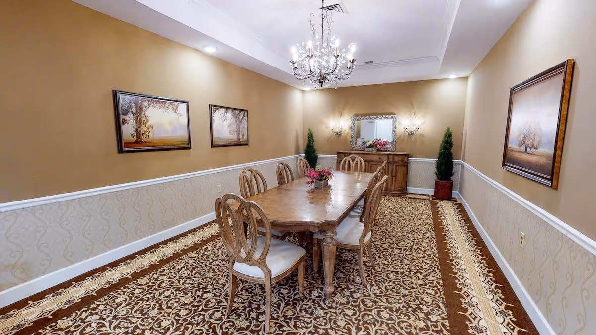 A formal dining room with a long wooden table surrounded by eight chairs with cushioned seats. The room features a patterned carpet, beige walls with white trim, three framed landscape paintings, two potted plants, a wooden sideboard with a decorative mirror above it, and a chandelier hanging from the ceiling.