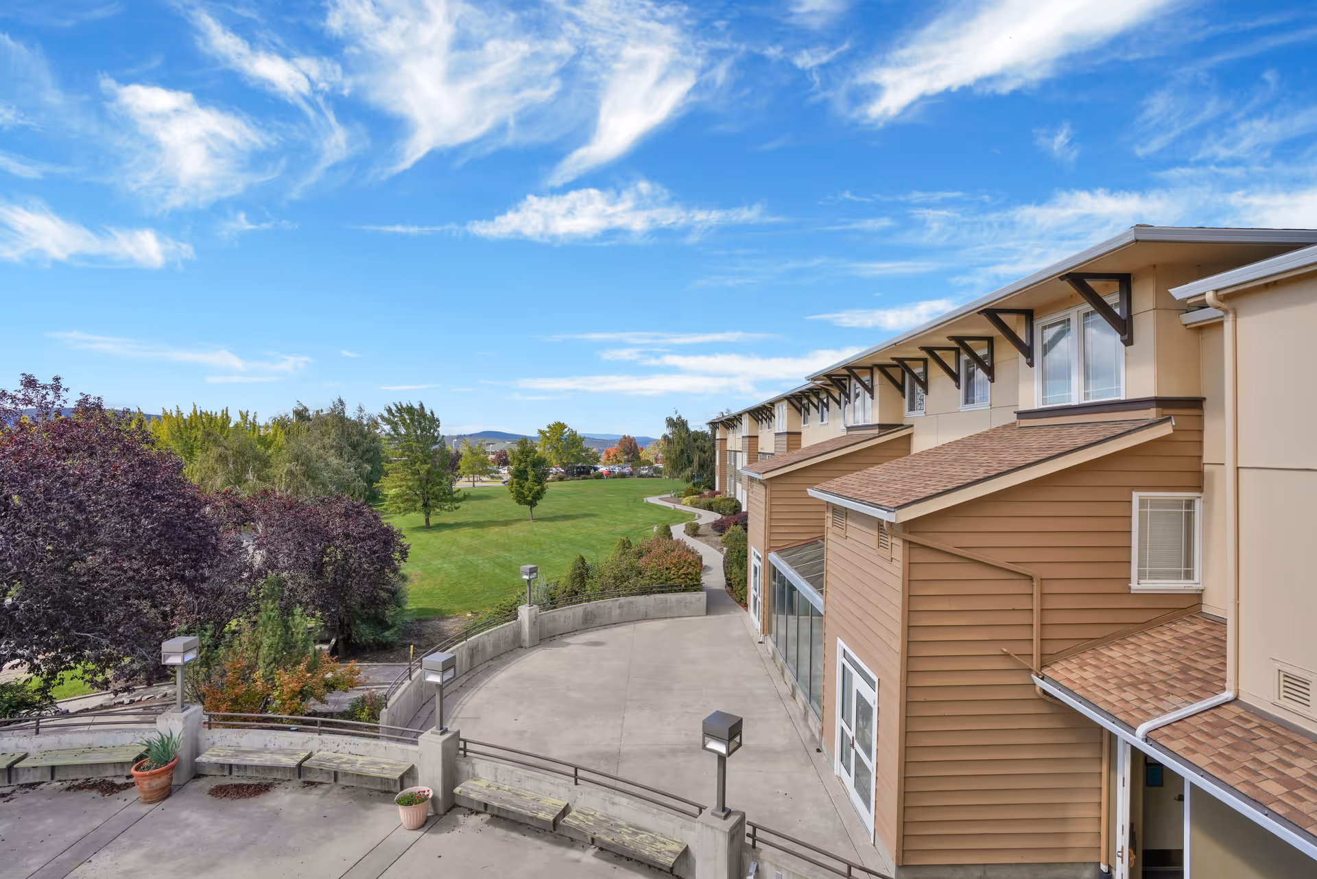 View of the exterior of a senior living facility building with beige and brown siding, adjacent to a large green lawn with trees under a partly cloudy blue sky.
