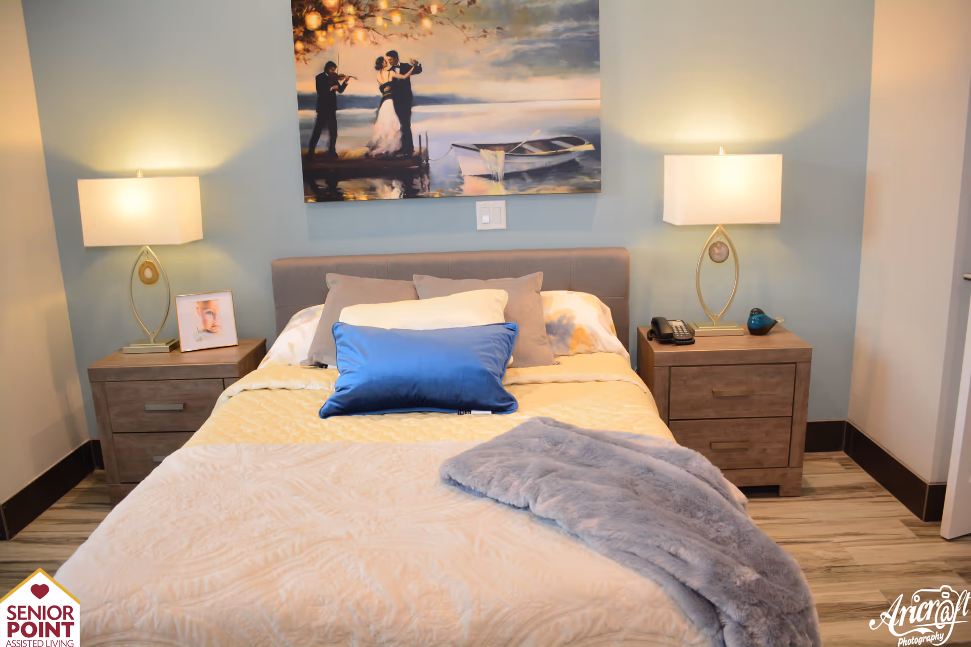 A neatly made bed with multiple pillows including a prominent blue one, flanked by two wooden nightstands each with a modern lamp. Above the bed hangs a painting of a couple dancing on a dock with a violinist playing beside them. The room has light blue walls and wooden flooring.