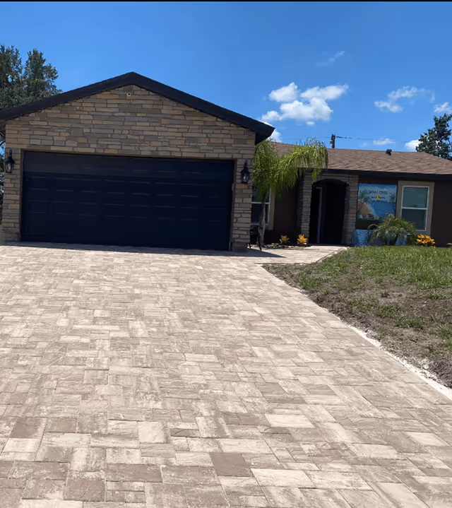 Front exterior view of a single-story house with a stone facade garage door, a paved driveway, and a small lawn area under a clear blue sky.