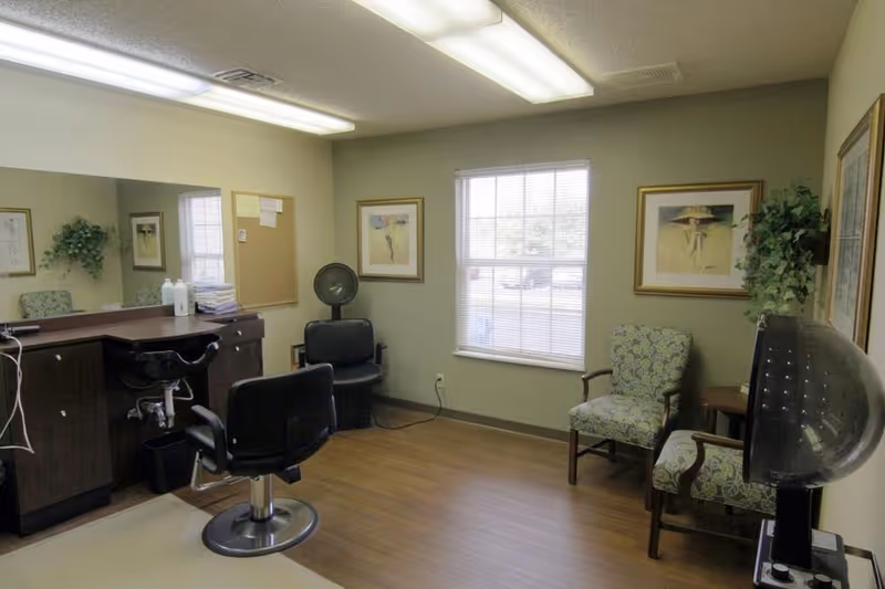 A small salon room with a styling chair in front of a counter with a large mirror. There is a hair dryer chair in the corner, two patterned armchairs near a window with blinds, framed artwork on the walls, and a potted plant on a small table.