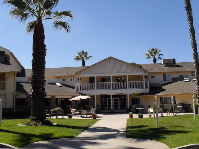 Exterior view of a senior living facility building with a two-story structure, a balcony, palm trees, outdoor seating with tables and umbrellas, and a paved walkway leading to the entrance under a clear blue sky.